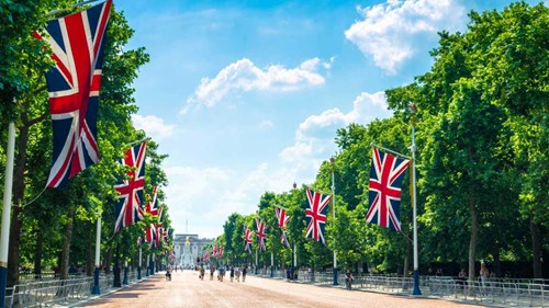 A photograph looking up The Mall in London towards Buckingham Palace, large Union Jack flags hang from flagpoles on either side of the street with green trees visible lining the street behind them. In the distance some figures walk up the street towards the Palace